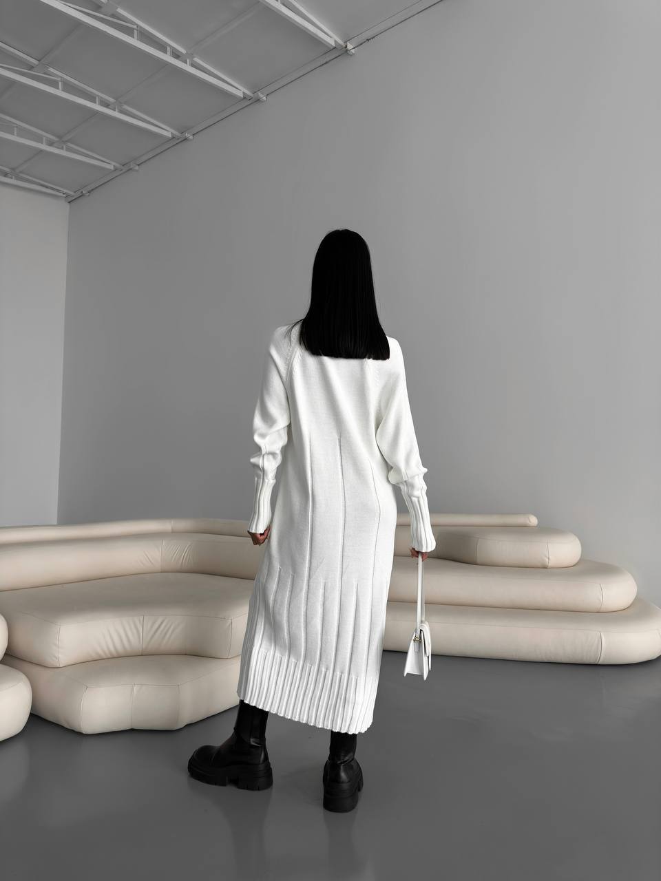 Person wearing a long white coat in a minimalistic room with gray walls and beige furniture.