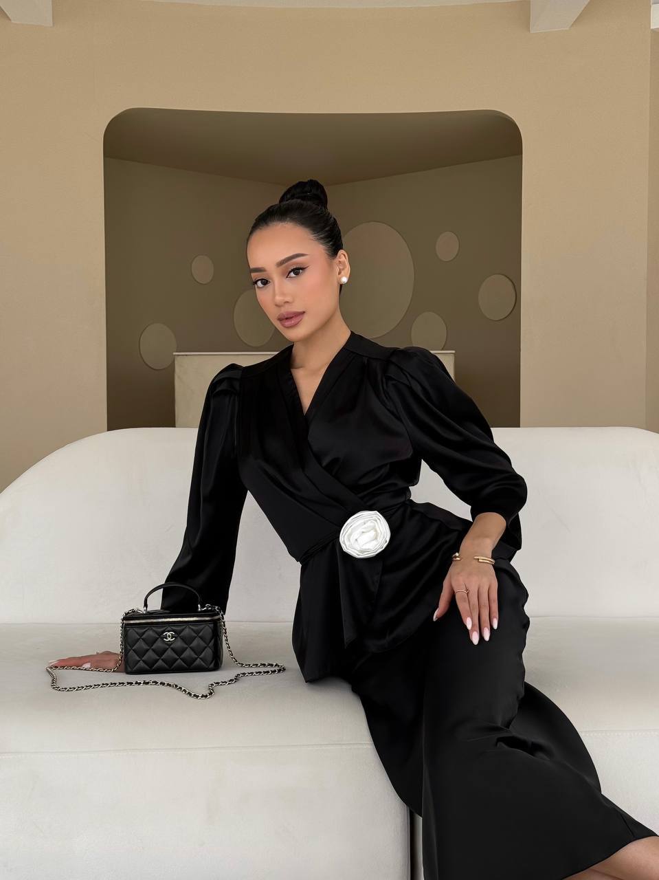 Woman in a black outfit sitting on a white couch with a black handbag.