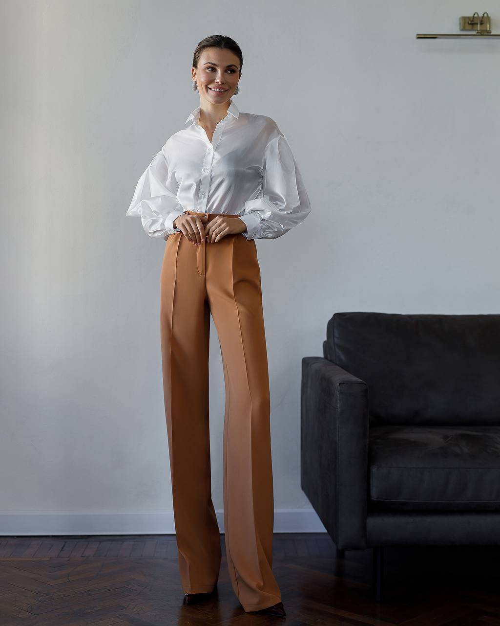 Woman wearing a white blouse and brown pants standing in a room with a gray wall and dark sofa.