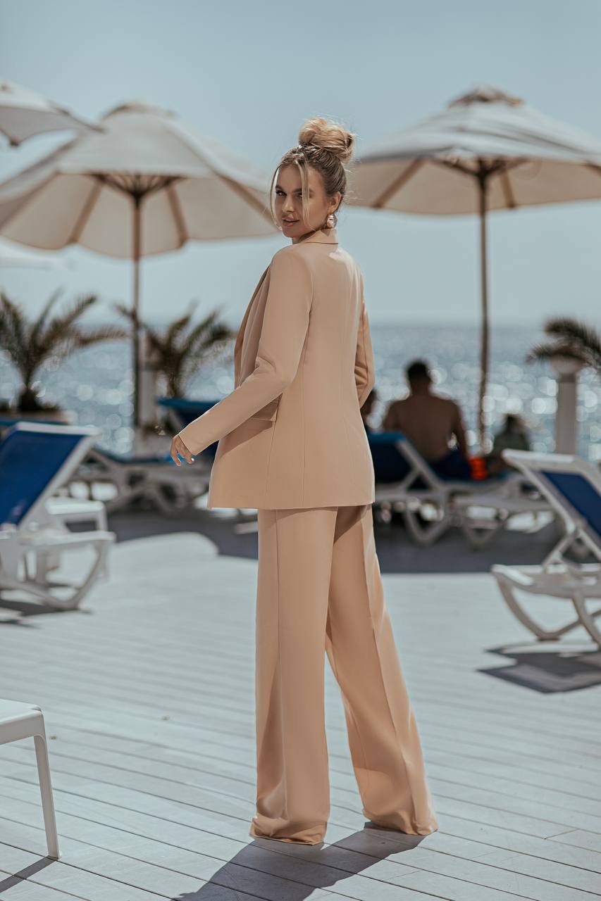 Woman in a beige suit standing on a beach deck with umbrellas and lounge chairs.
