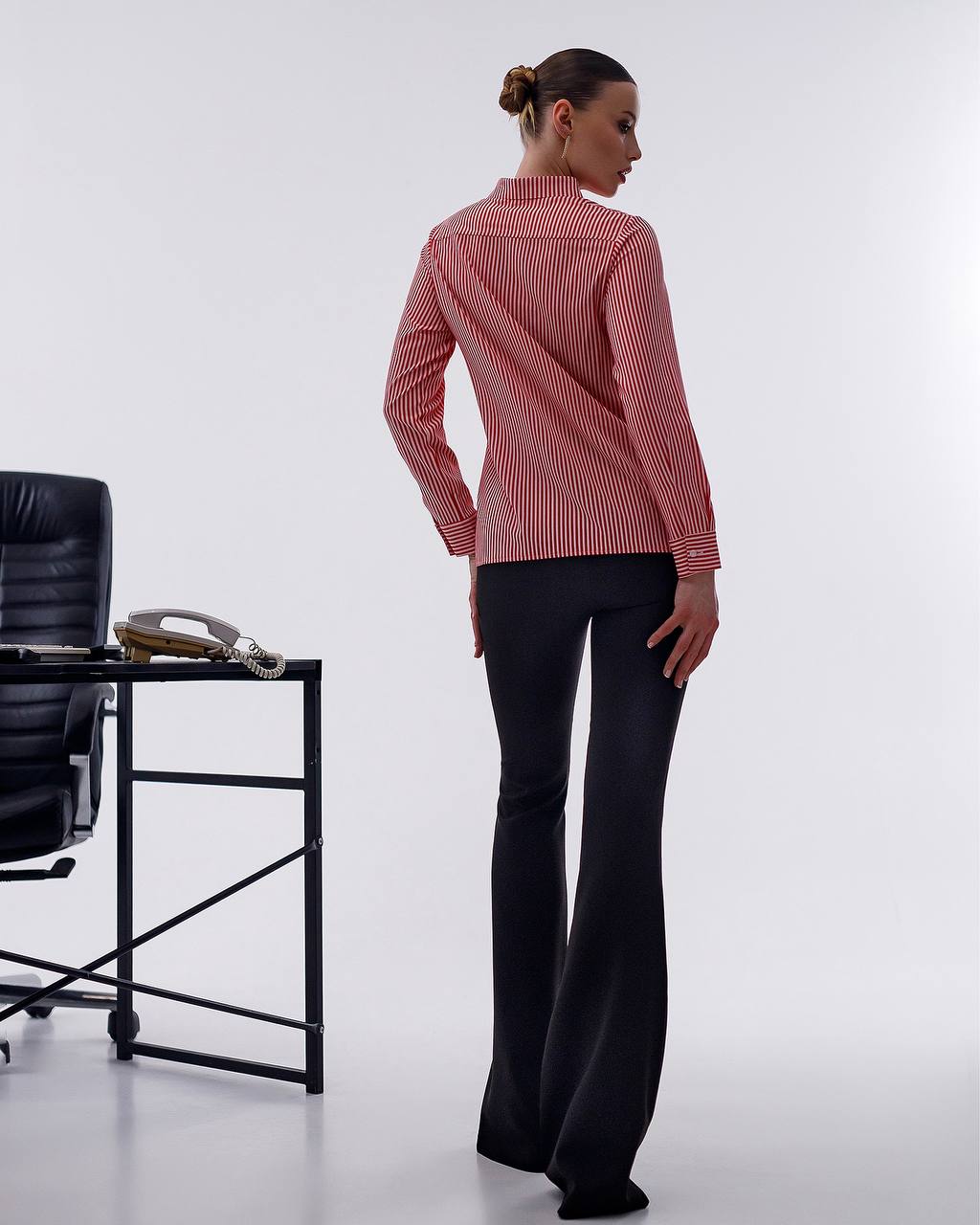 Woman wearing a red and white striped shirt and black pants standing next to a black office chair on a white background