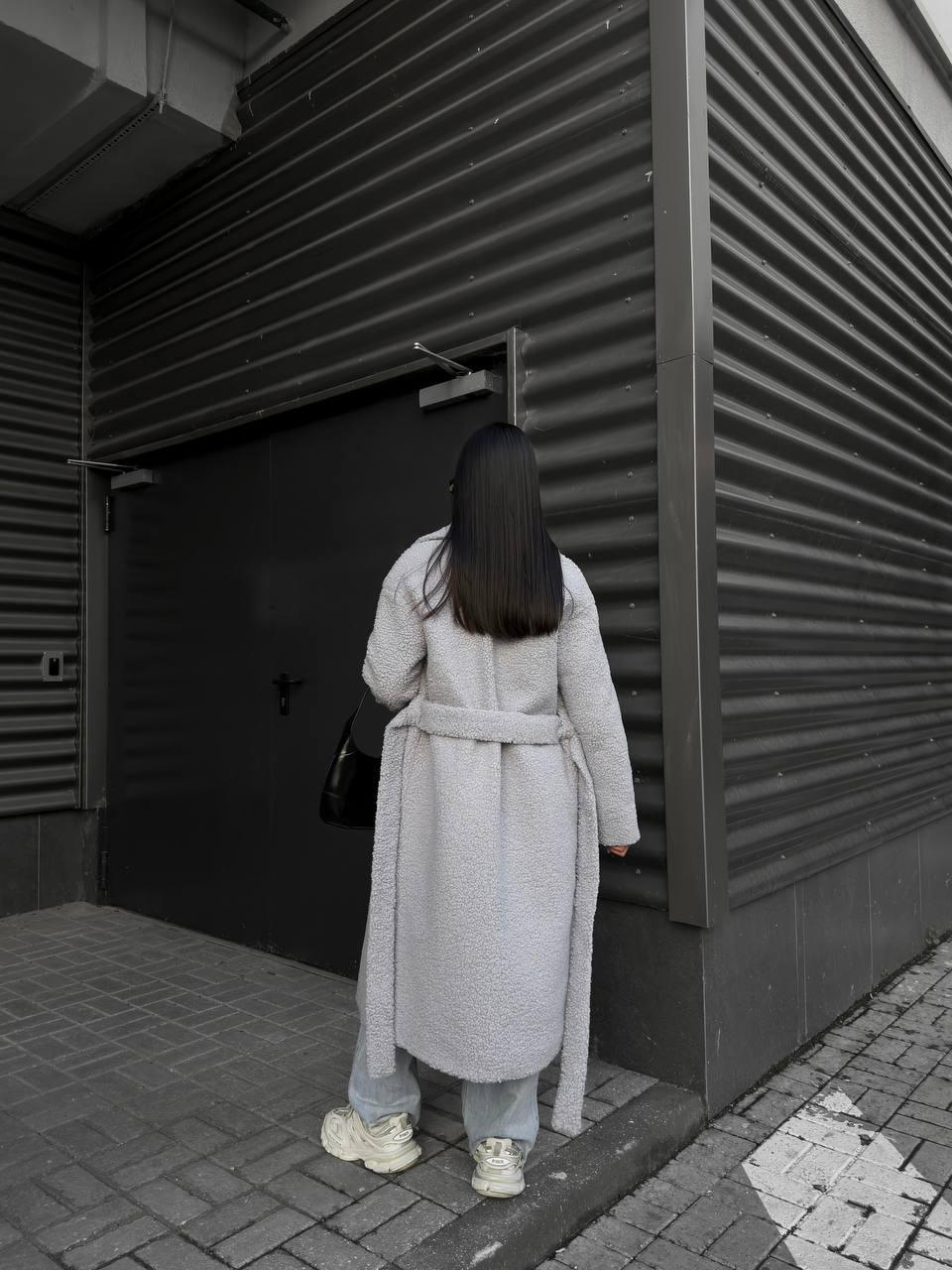 Person in a long coat standing in front of a dark door with horizontal slats.