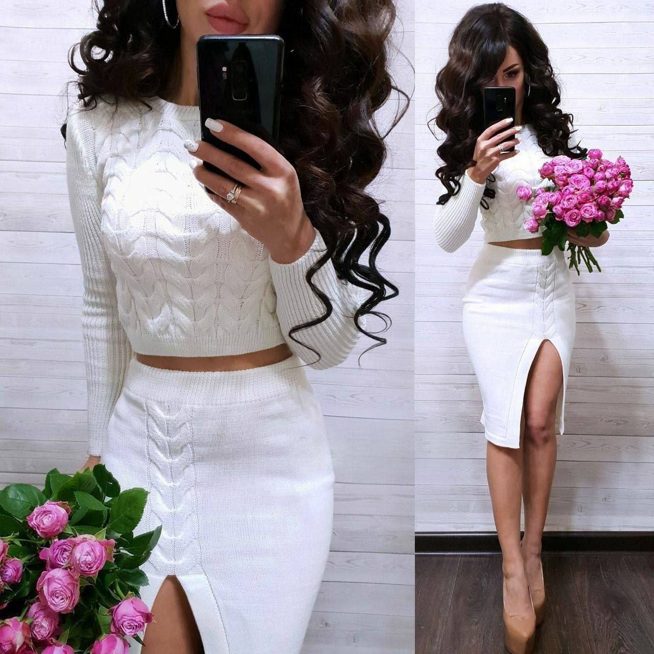 Woman in a white outfit with a high slit, holding pink flowers, against a white wooden panel background.