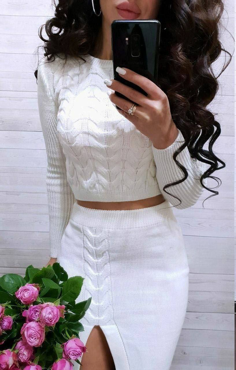 Person wearing a white cable knit sweater and skirt with a phone in front of a white wooden wall.