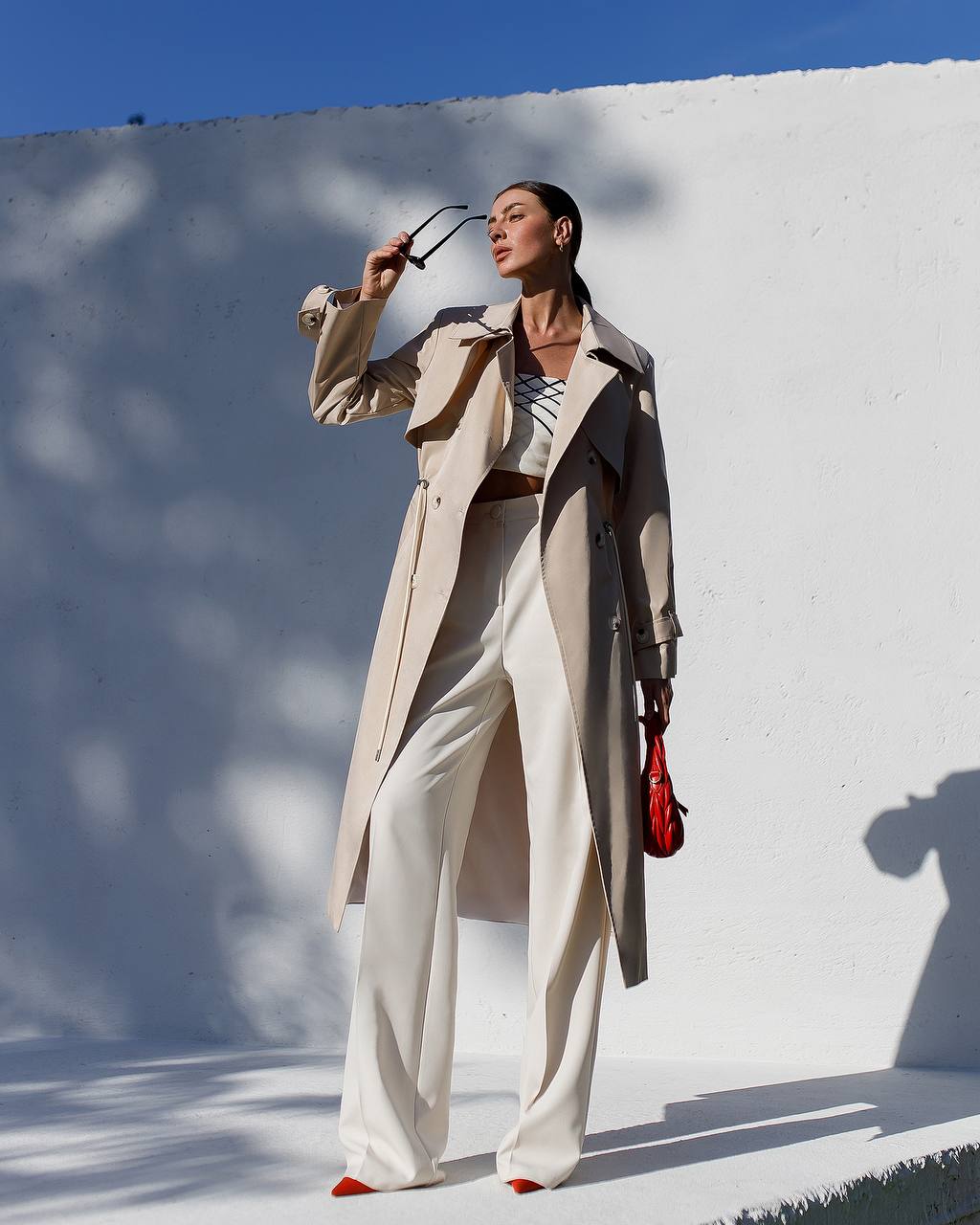 Woman in a beige trench coat and white pants standing against a white wall with blue sky.