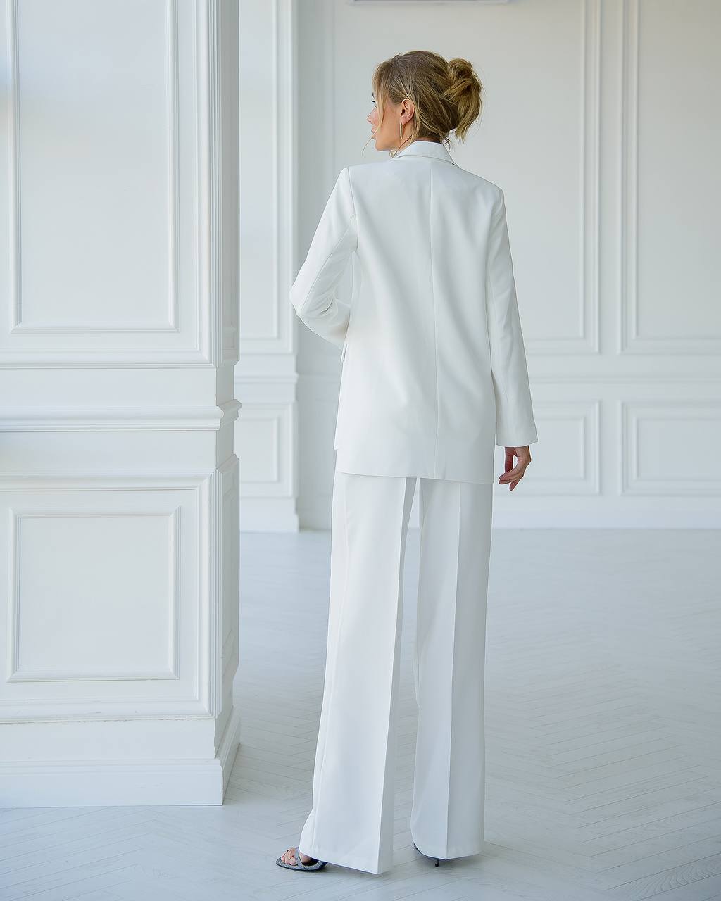 Woman wearing a white suit standing in a minimalistic room with white walls.