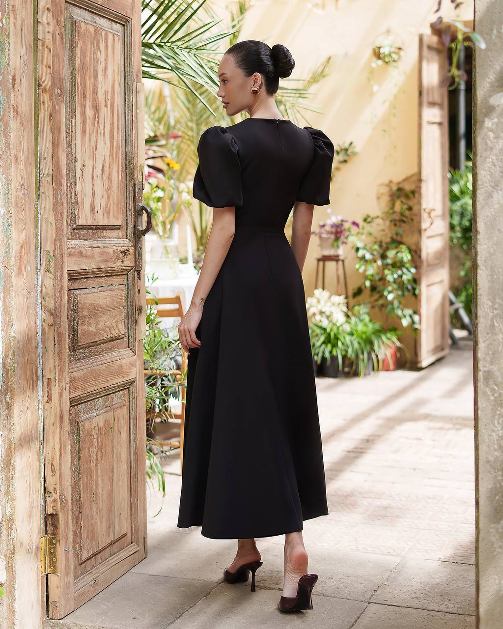 Woman in a black dress standing in a sunlit outdoor area with wooden doors and plants.