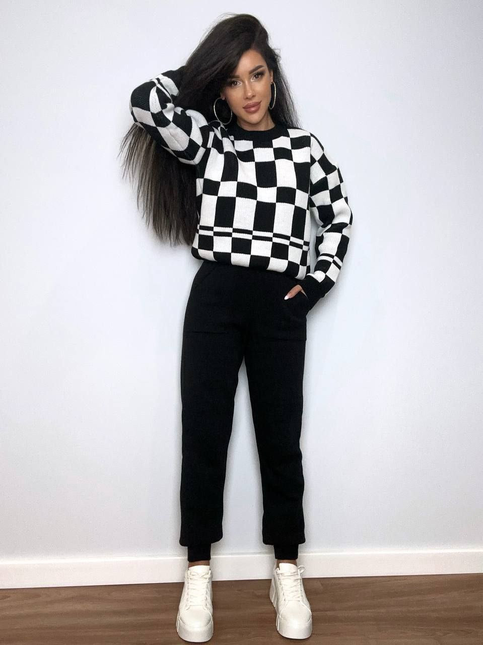 Person wearing a black and white checkered sweater with black pants and white shoes against a white wall.
