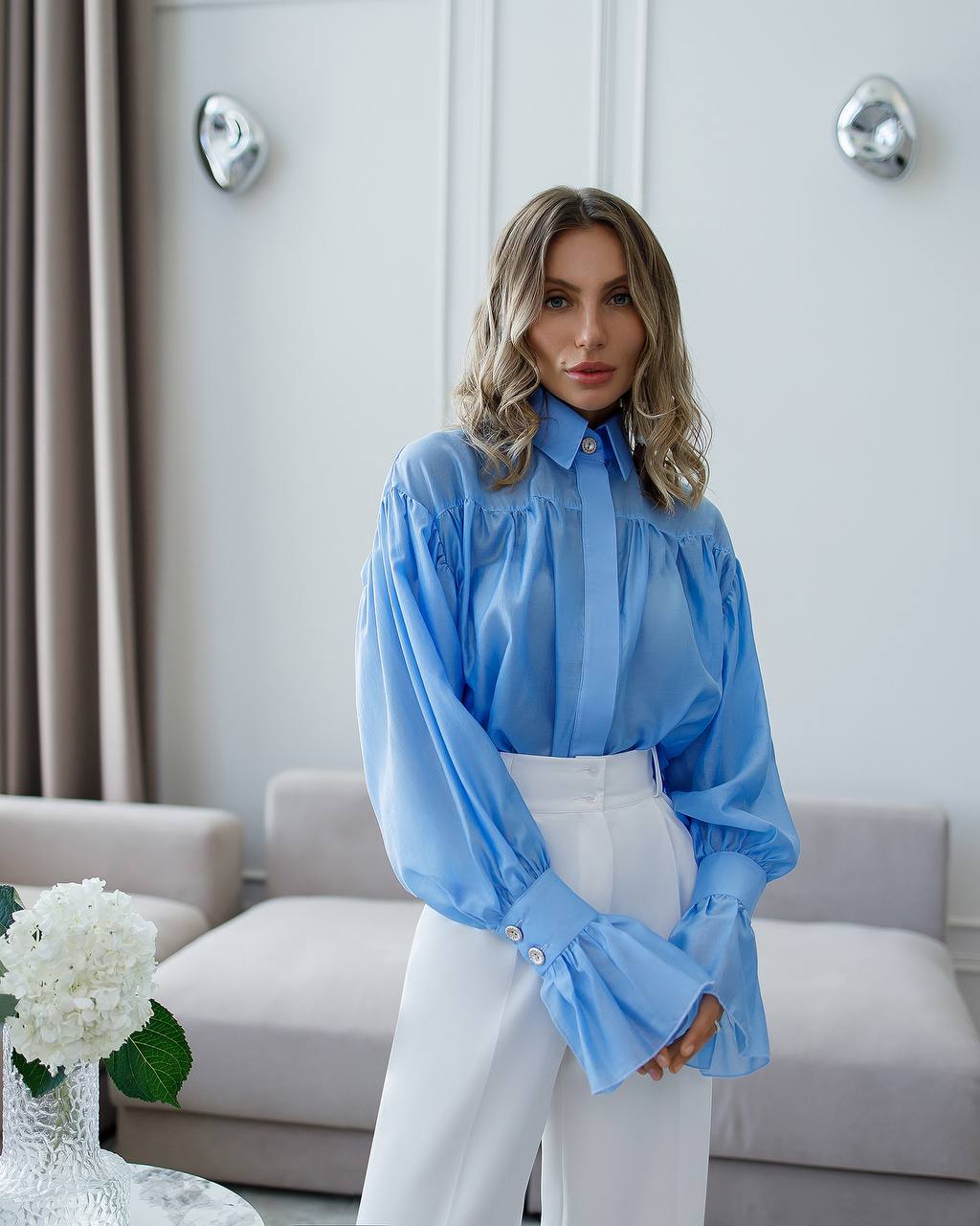 Woman wearing a blue blouse and white pants in a modern living room.