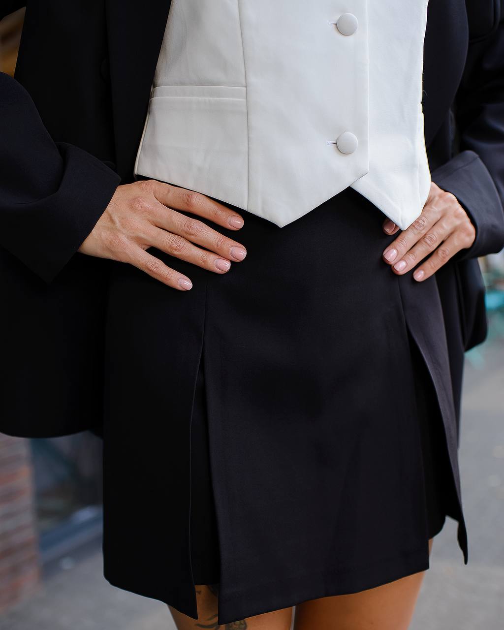 Person wearing a black blazer, white vest, and black skirt with hands on hips.