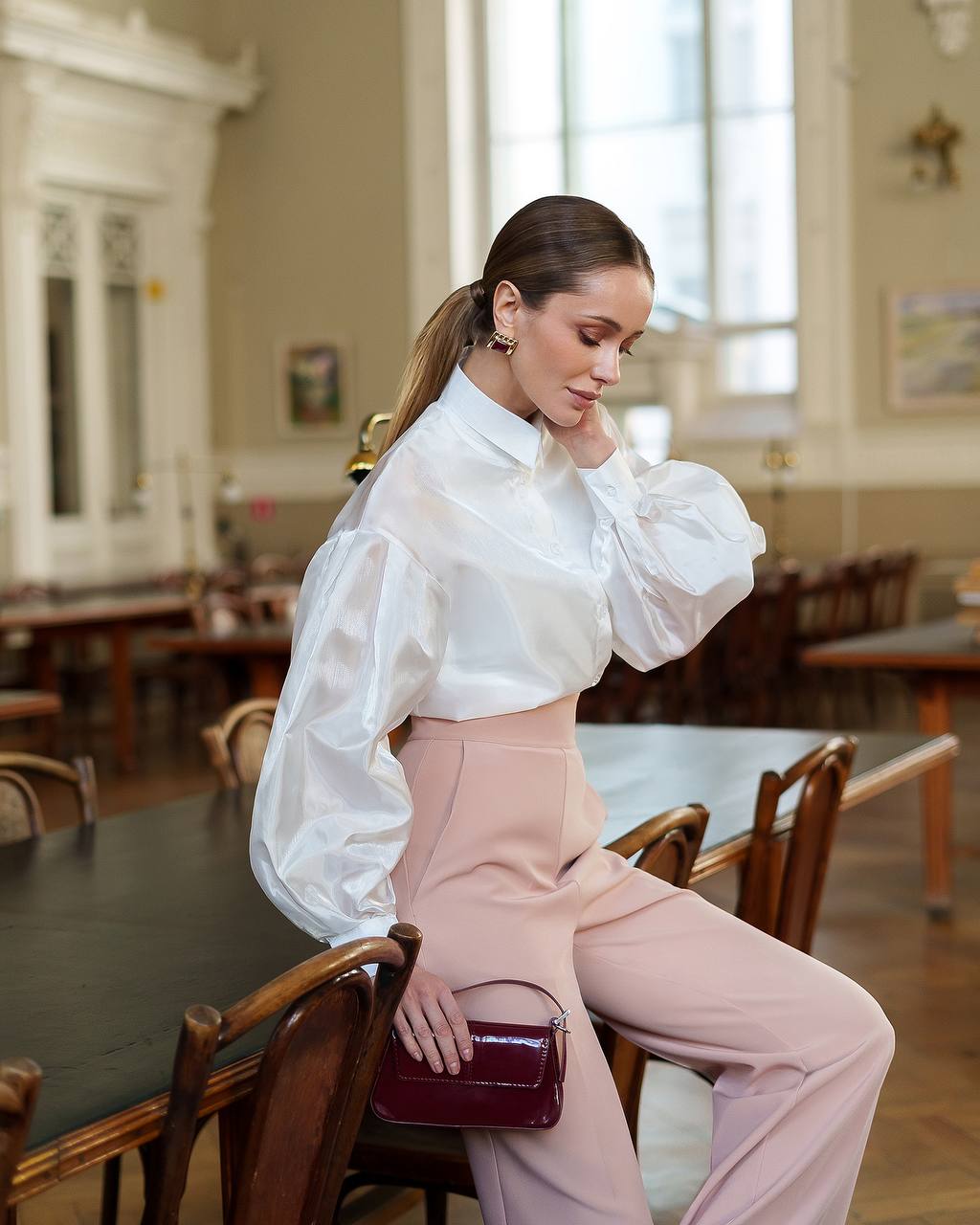 Woman in a white blouse and pink pants sitting at a table in a room with large windows.
