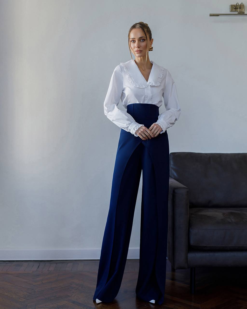 Woman wearing a white blouse and blue pants standing in a room with a gray sofa.