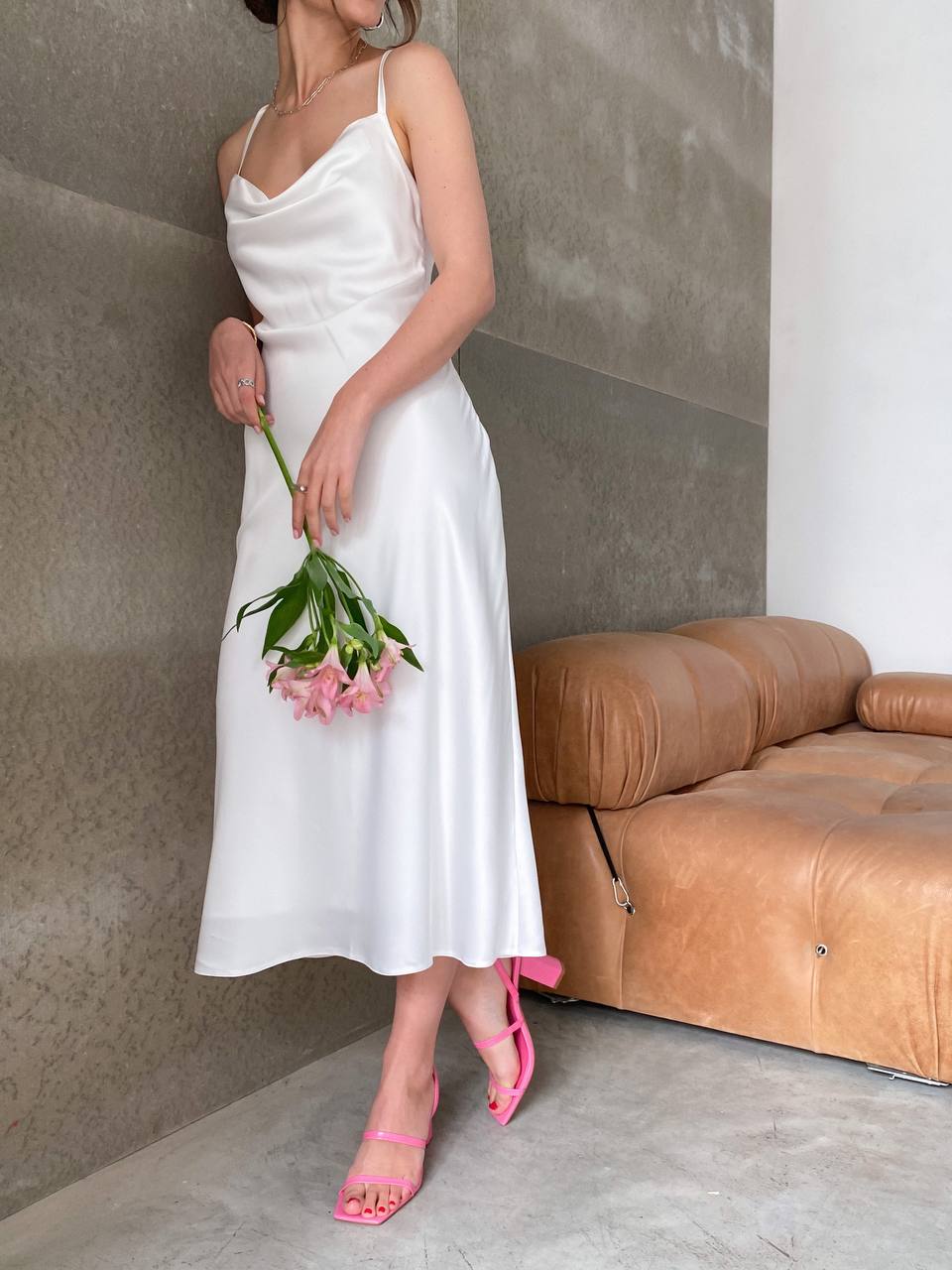Woman in a white dress holding pink flowers in a room with a brown sofa.