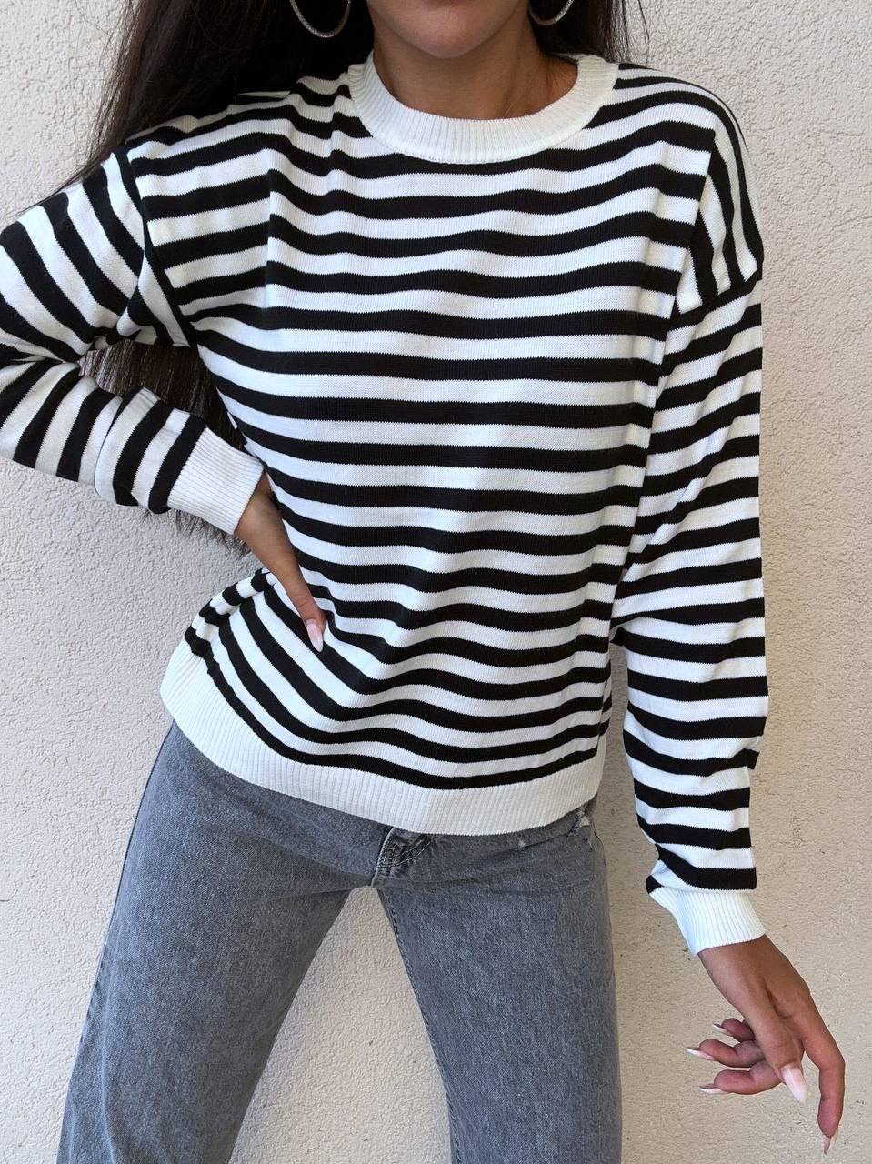 Person wearing a black and white striped sweater with gray jeans against a beige wall.
