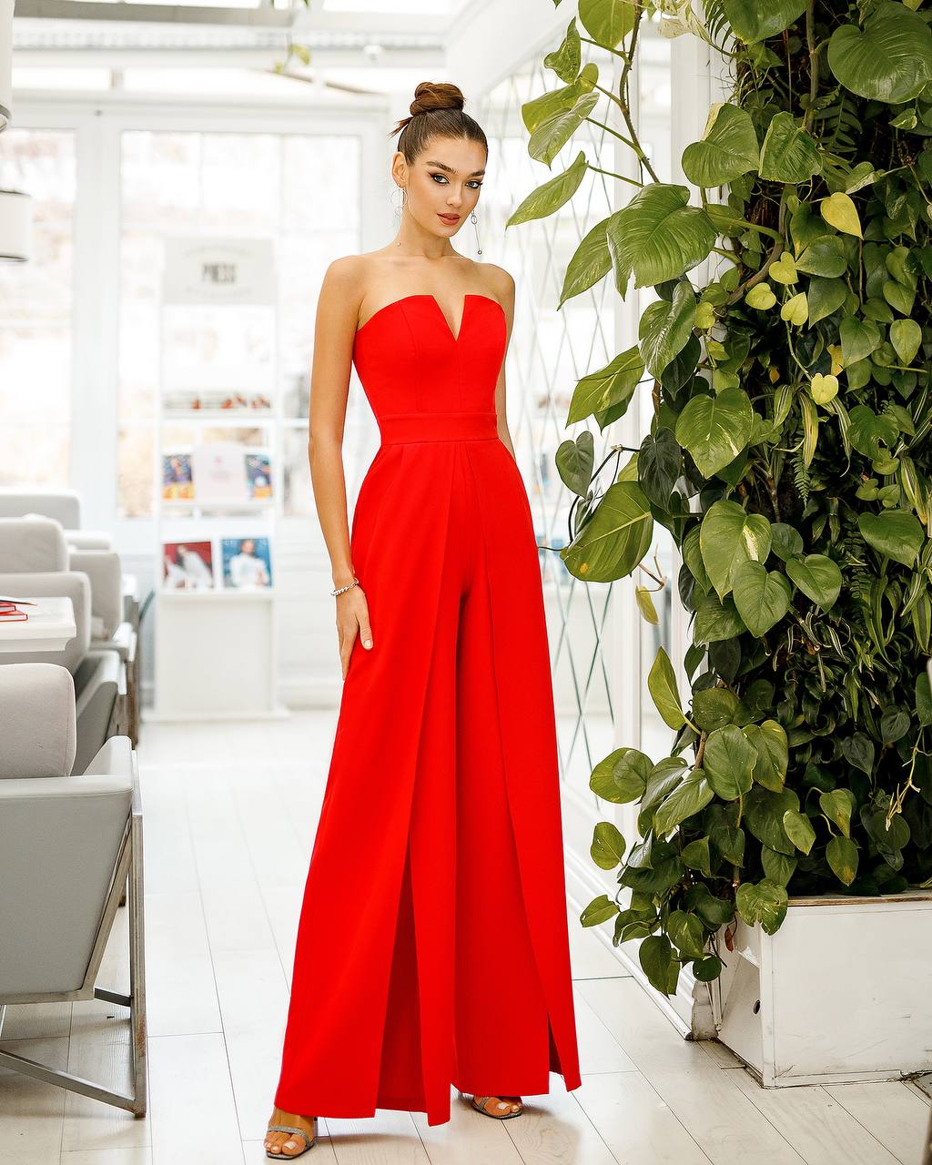 Woman in a red strapless jumpsuit standing indoors with plants and furniture in the background