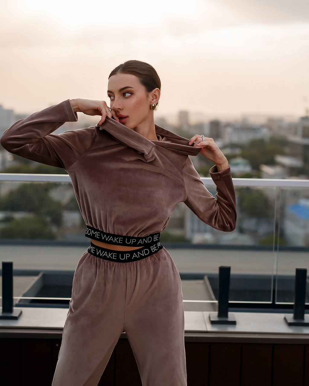 Woman in a brown velvet outfit with a black belt standing on a balcony with a cityscape view.