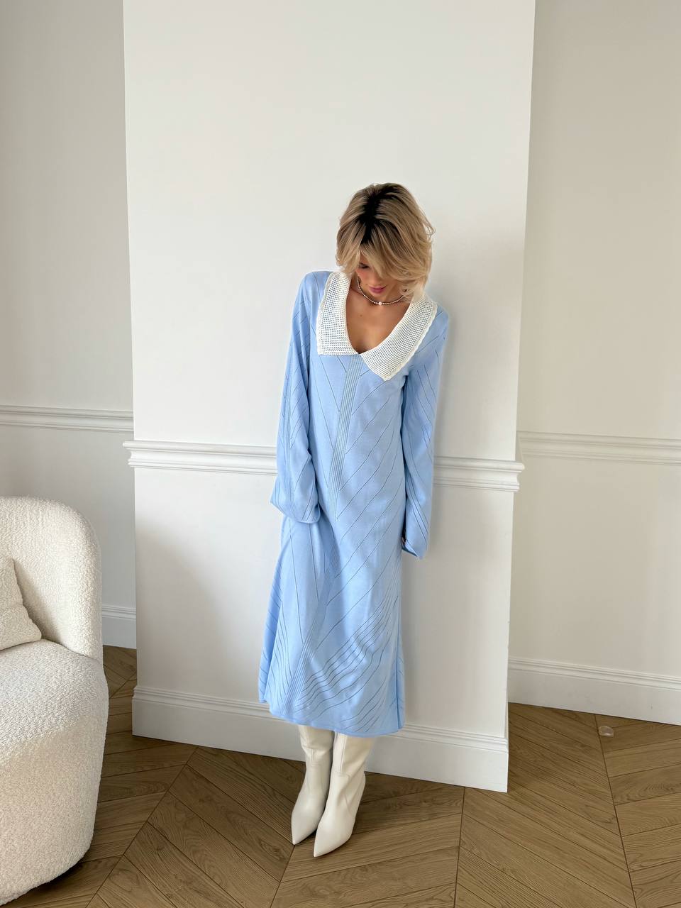 Person wearing a light blue dress with a white collar in a room with a white wall and wooden floor.