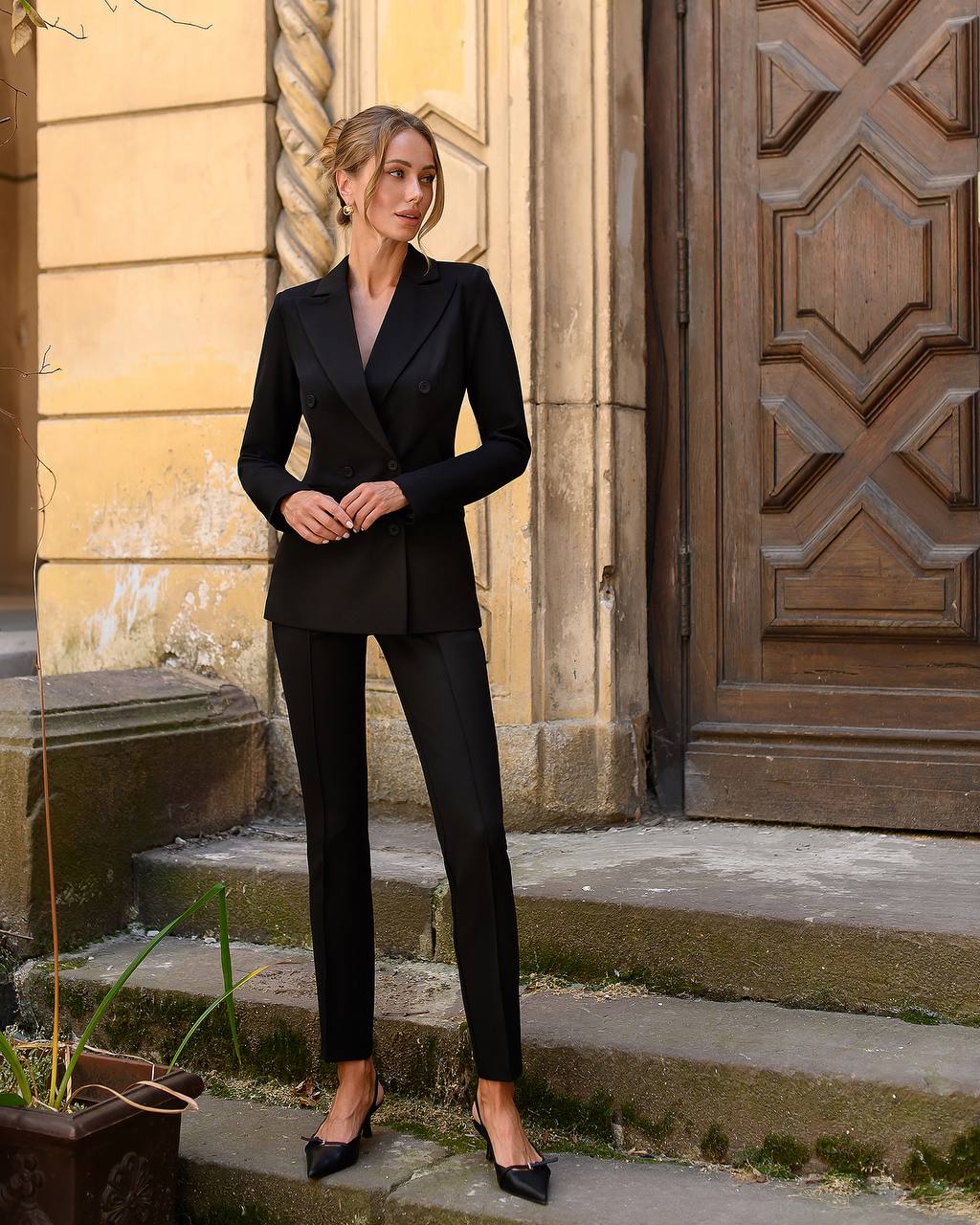 Woman in a black suit standing on steps in front of a wooden door.