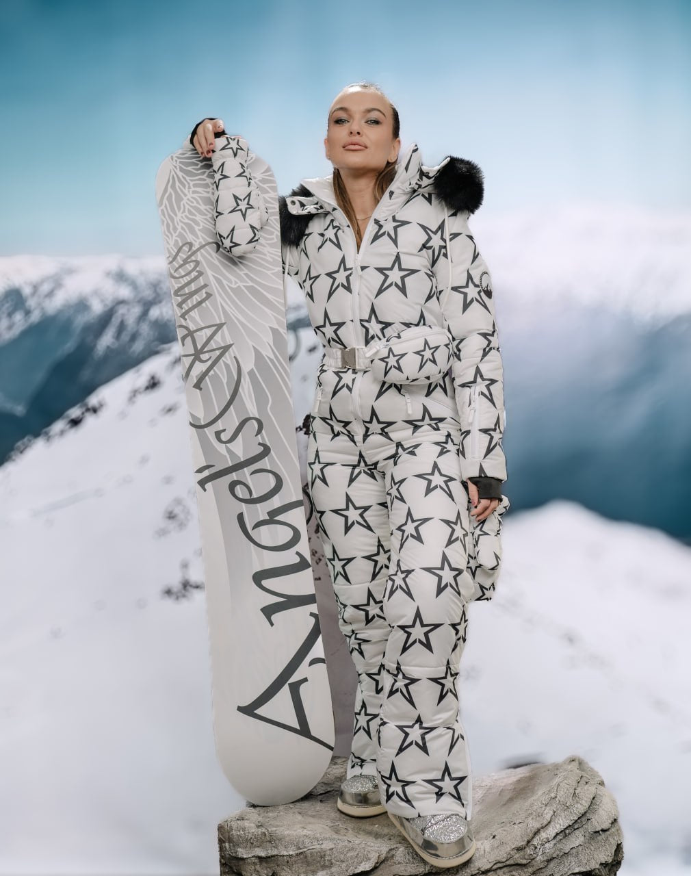 Person in star-patterned snowsuit holding a snowboard with mountains in the background