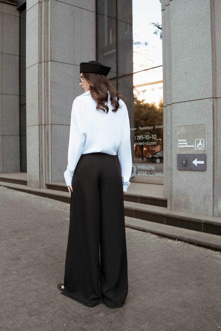 Person wearing a white shirt and black wide-leg pants standing in front of a building entrance.