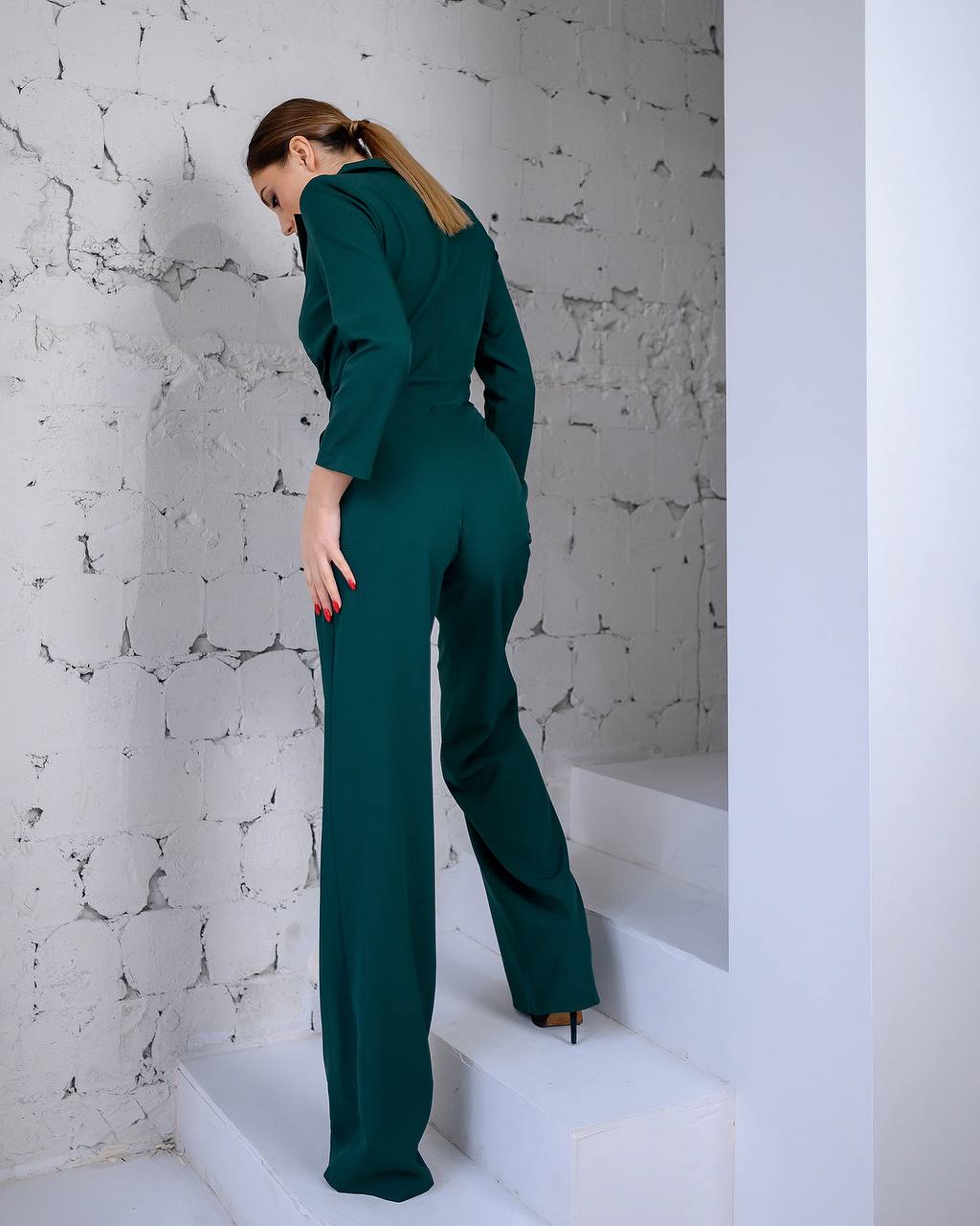 Woman in a green jumpsuit standing on white steps against a white marble wall.