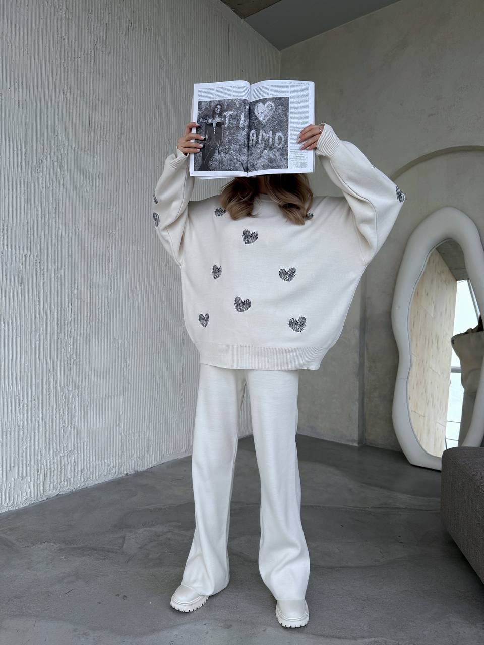 Person wearing a white sweater with heart patterns and white pants, holding a book in front of their face.