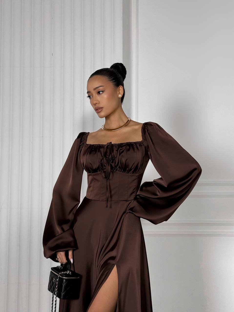 Woman wearing a brown dress with long sleeves against a white wall.