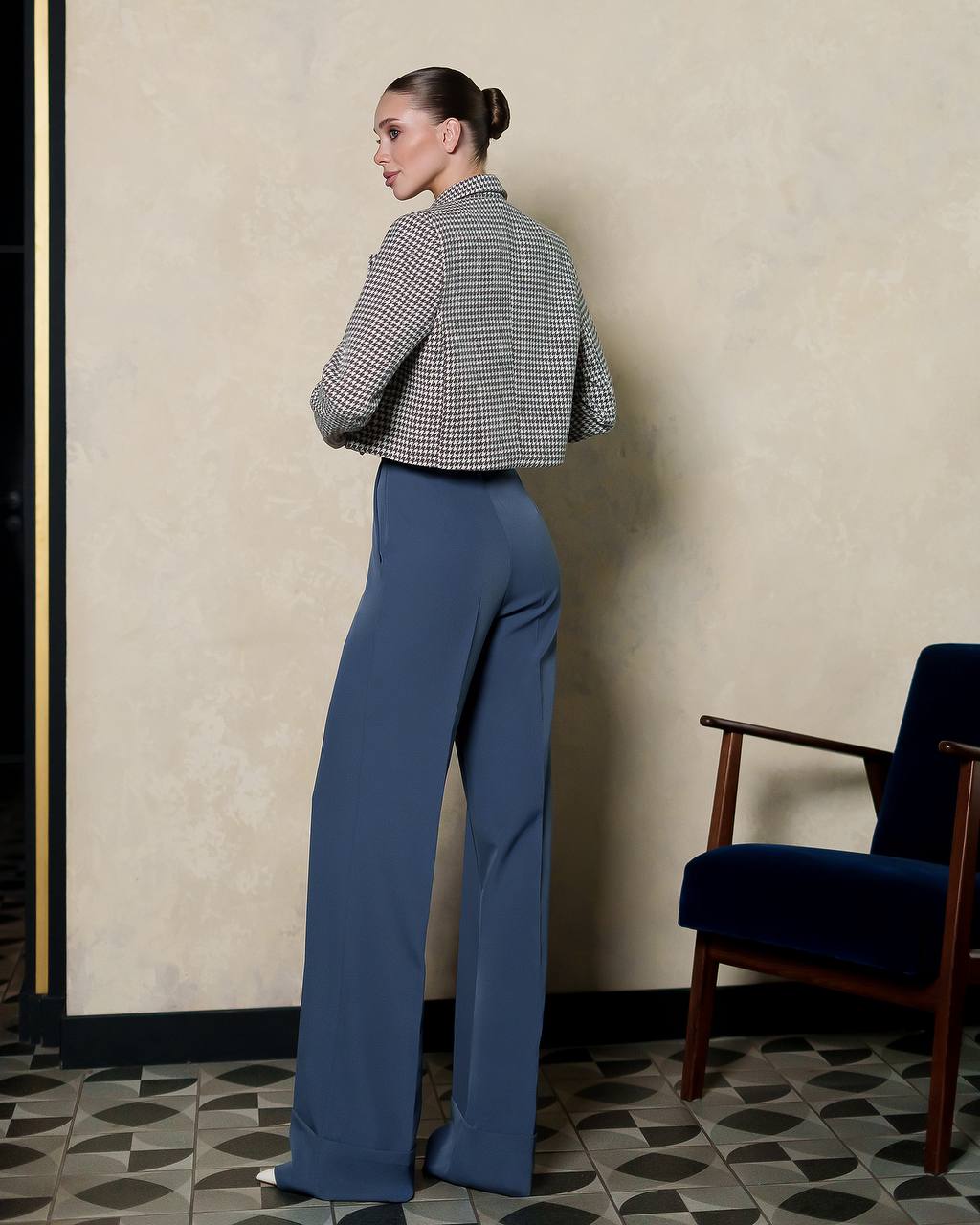 Woman in a checkered jacket and blue pants standing against a beige wall with a chair in the corner.