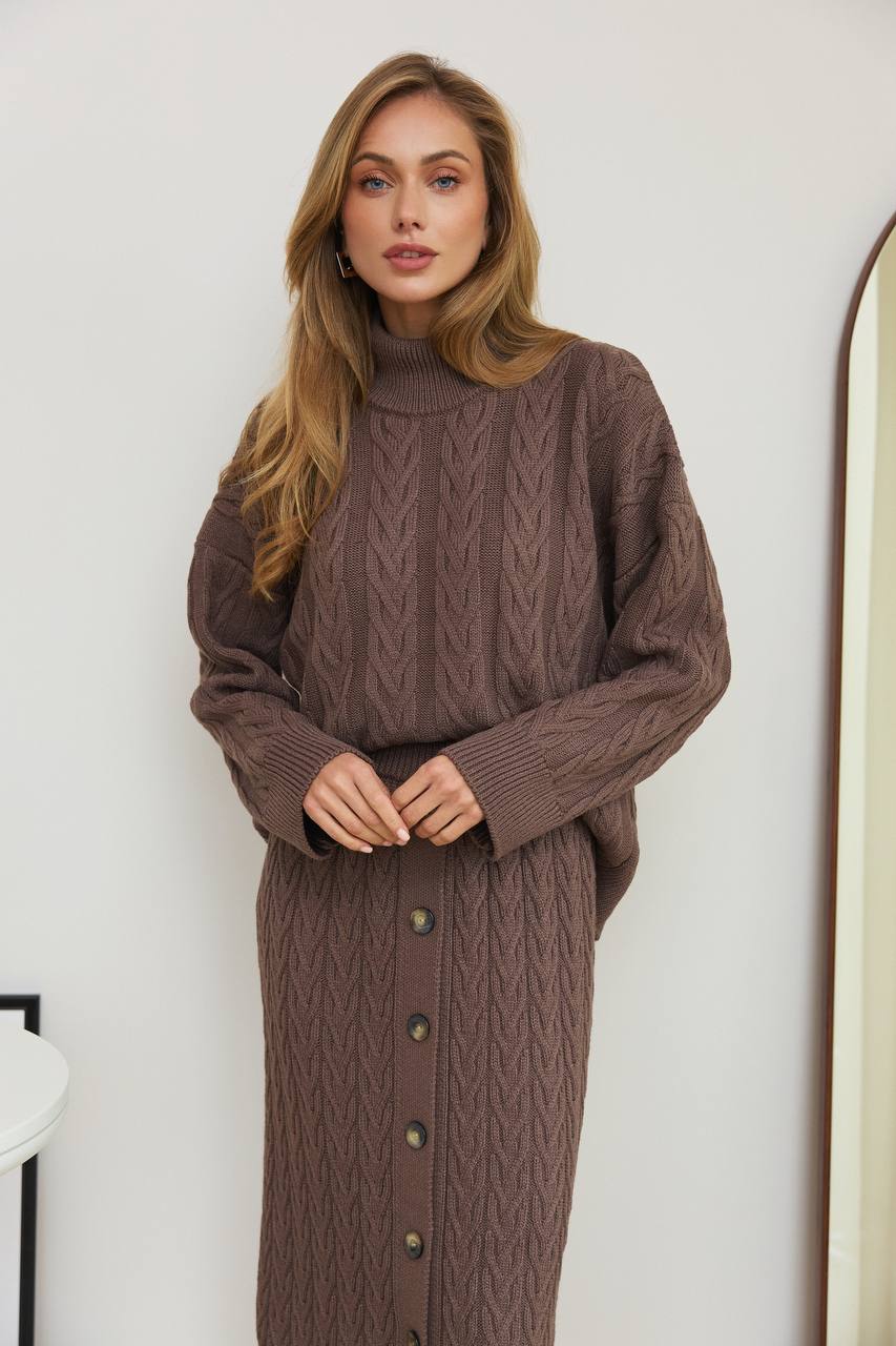 Woman wearing a brown cable knit sweater dress against a white wall.