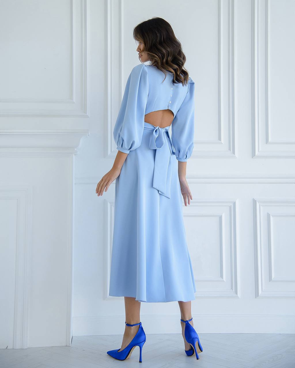 Woman wearing a light blue dress with cut-out design and matching shoes against a white paneled wall.