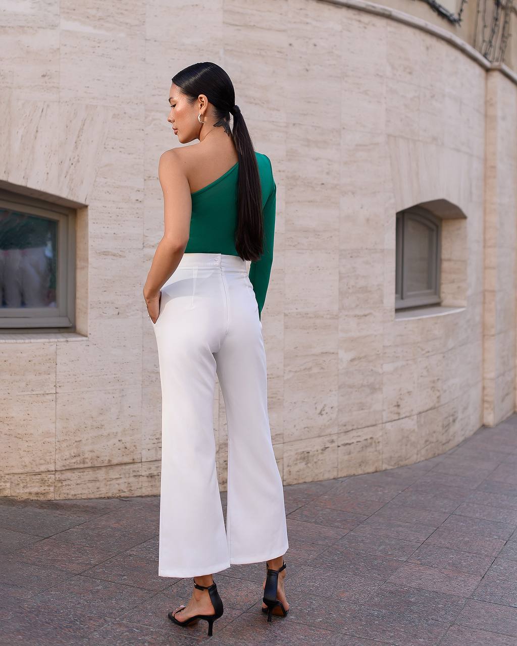 Woman in green top and white pants standing against a beige building.