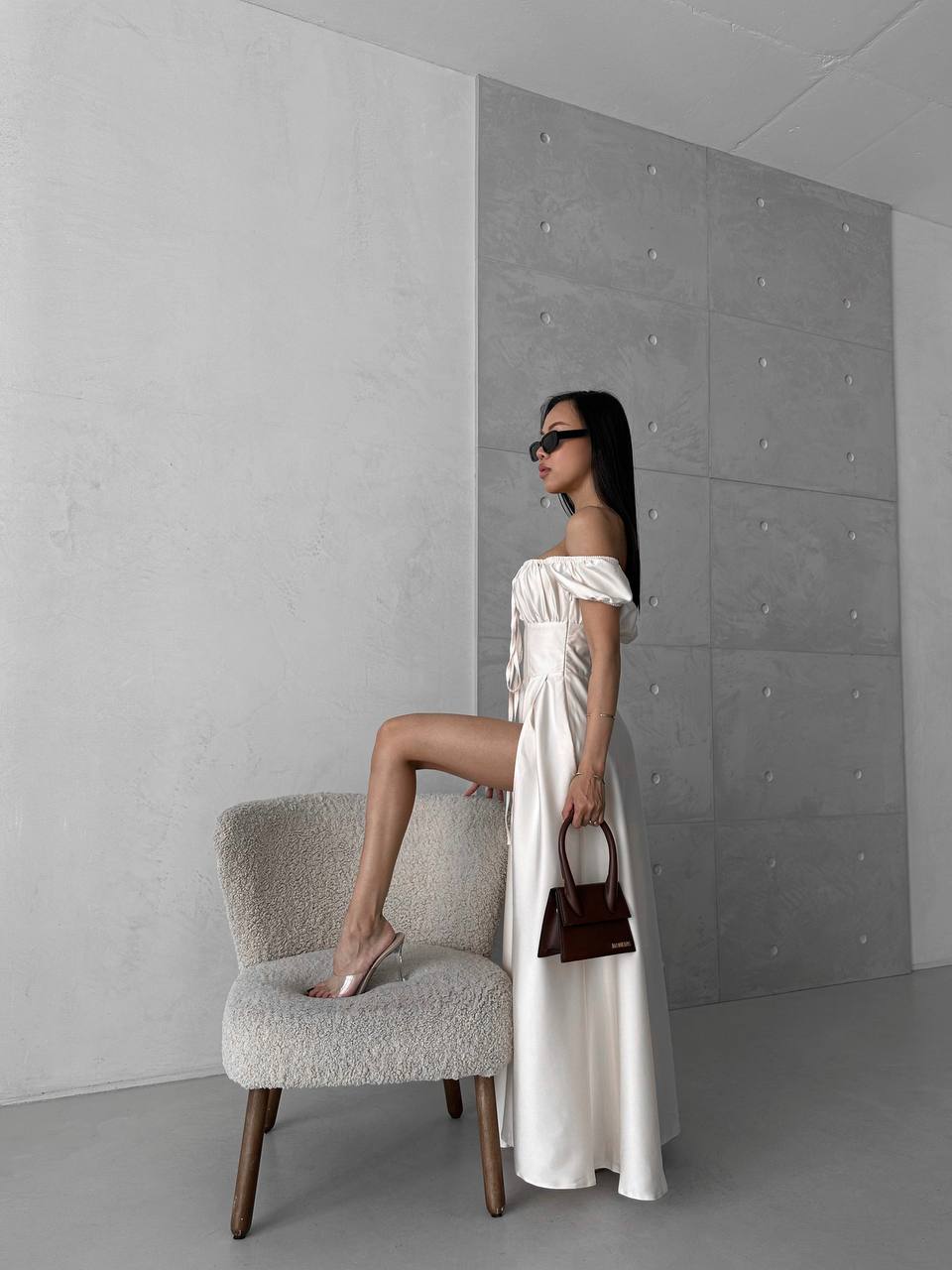 Woman in a white off-shoulder dress standing next to a gray chair against a gray wall.