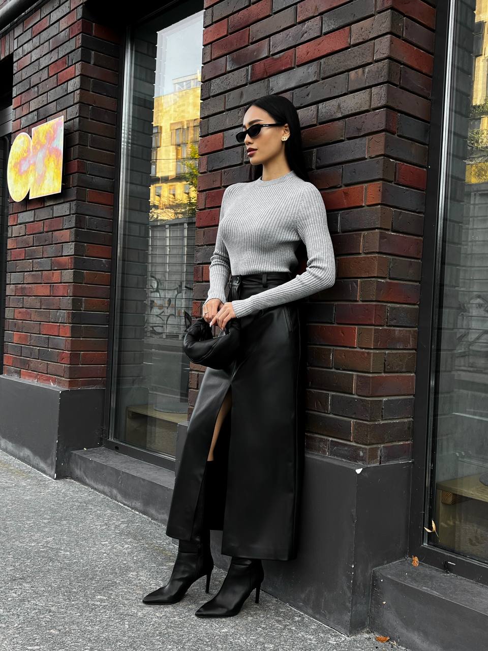 Woman in a gray sweater and black skirt leaning against a brick wall.