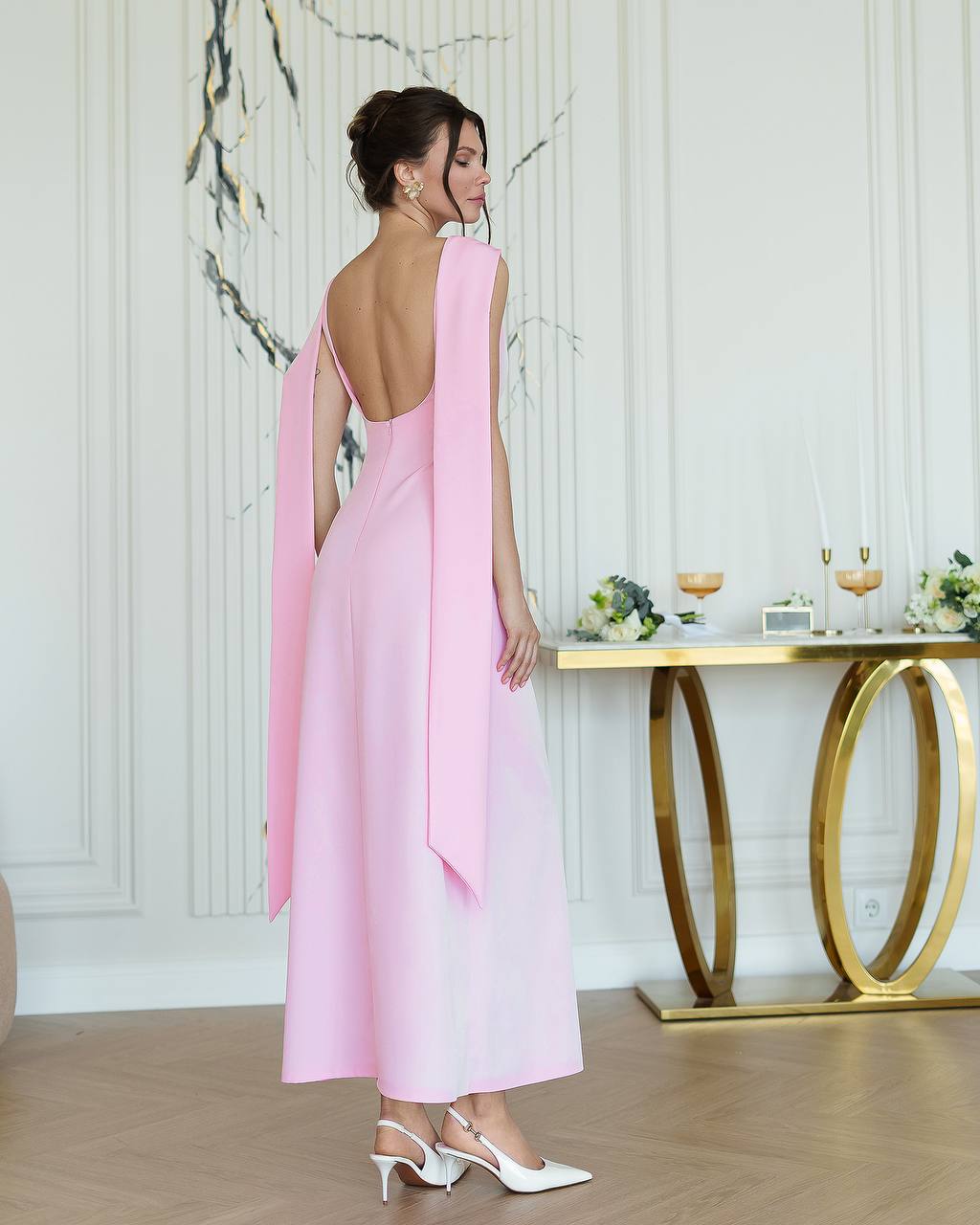 Woman in a pink dress with a backless design standing in a room with a decorative table.
