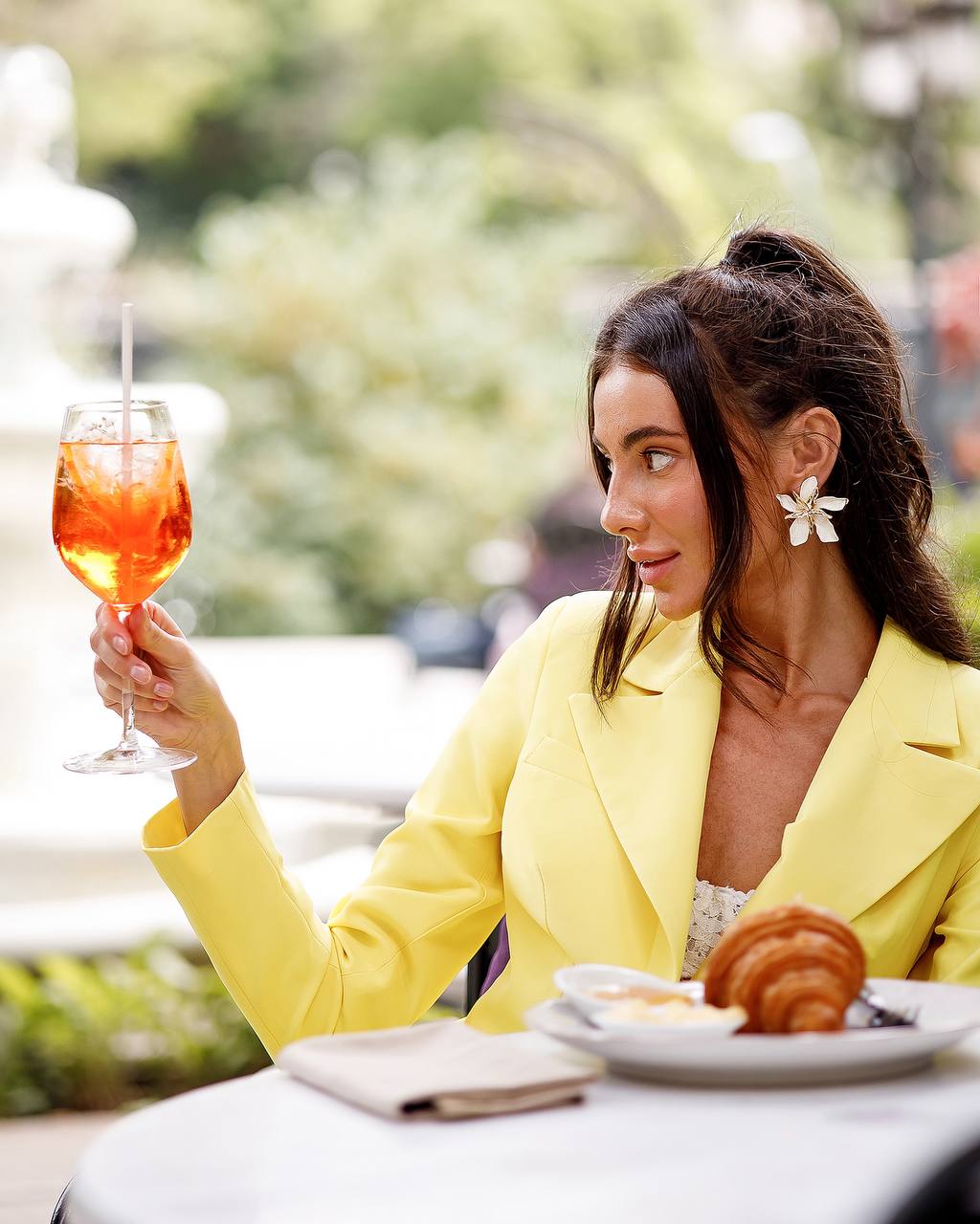 Woman in a yellow blazer holding a glass of orange cocktail outdoors