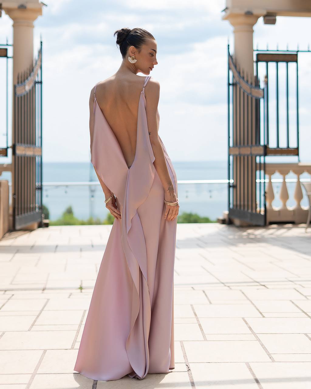 Woman in a pink evening gown with a backless design standing on a balcony with ocean view.