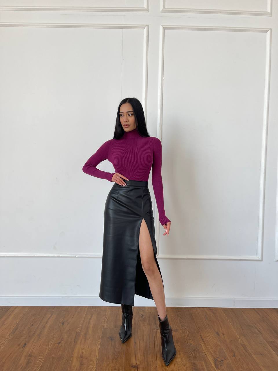 Woman wearing a purple turtleneck and black leather skirt against a white paneled wall.