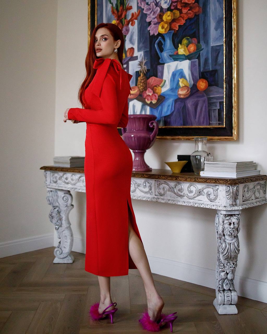 Woman in a red dress standing in a room with decorative elements and a painting on the wall.
