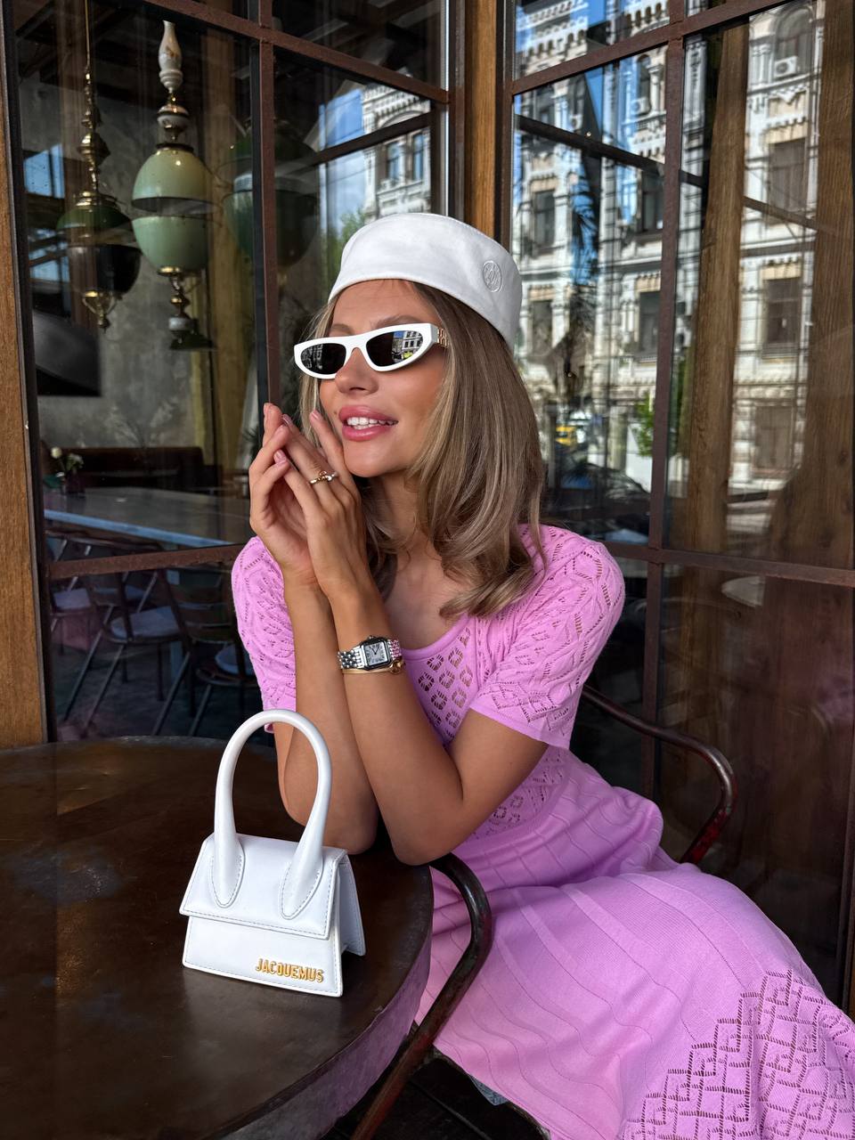 Woman in a pink dress and white cap sitting at a table with a small white handbag.