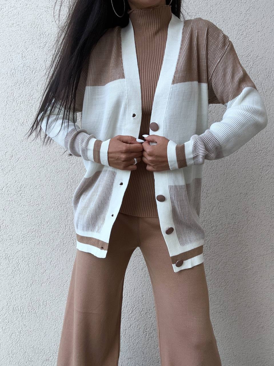 Person wearing a brown turtleneck, white cardigan with buttons, and brown pants against a plain background