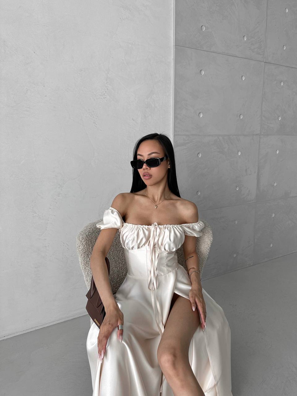 Woman in a white off-shoulder dress sitting on a chair against a gray wall.