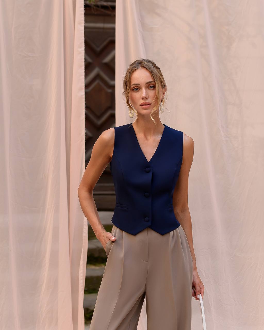 Woman wearing a navy blue vest and beige pants standing in front of a beige curtain.