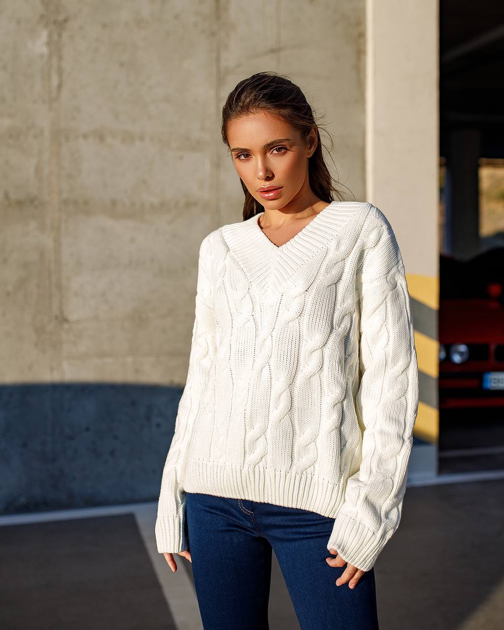 Woman wearing a white cable knit sweater and blue jeans standing against a concrete wall.