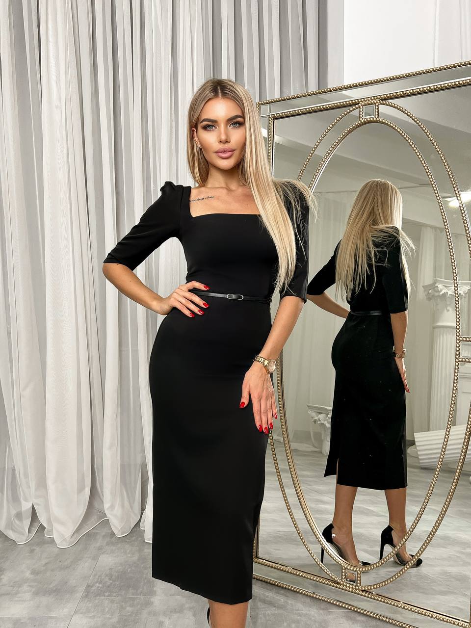 Woman in a black dress standing in front of a mirror in a room with white curtains.