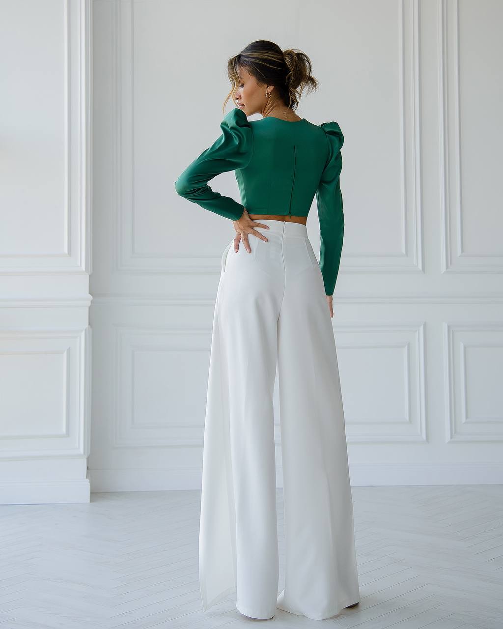 Woman wearing a green top and white pants in a white room