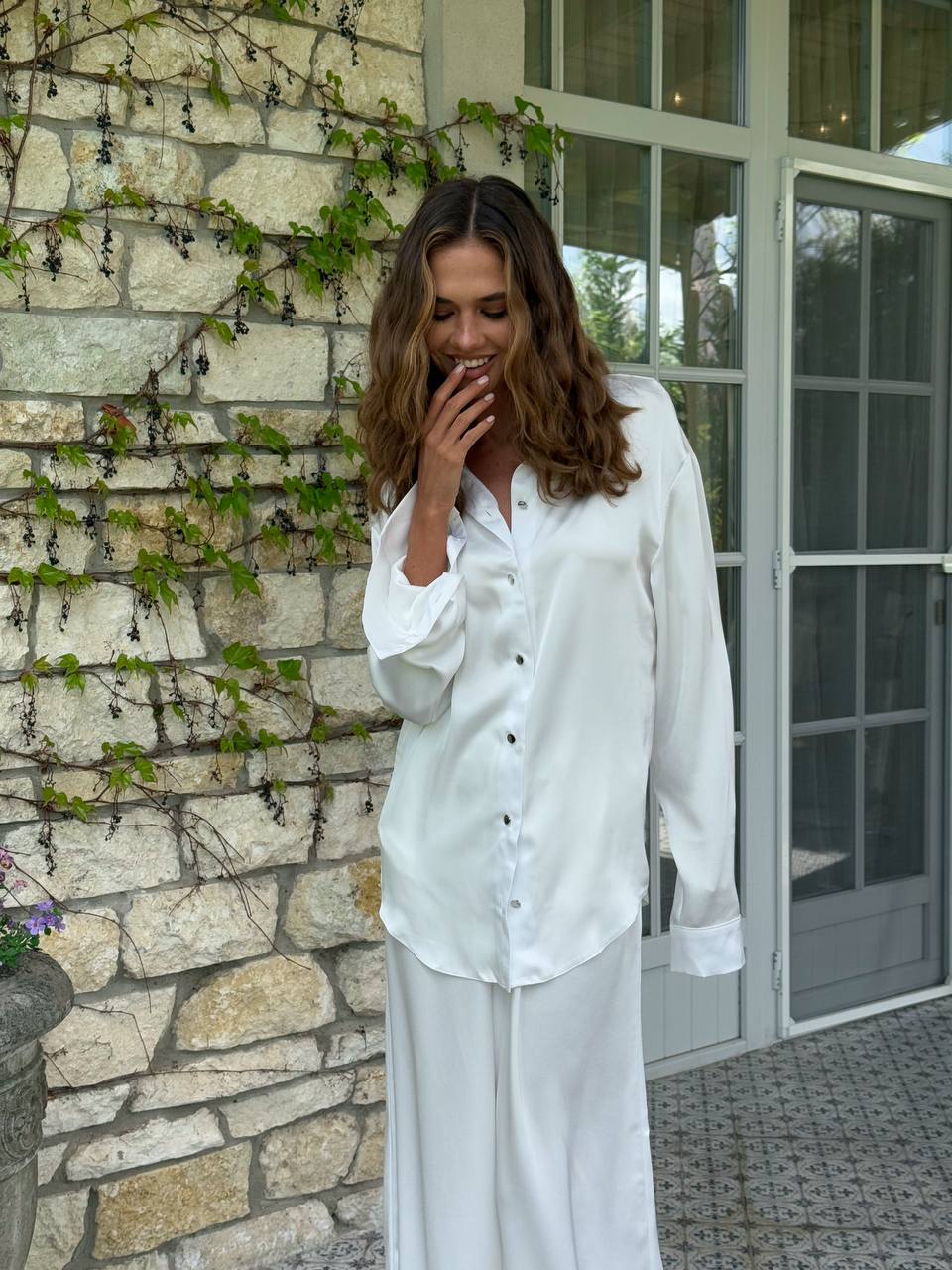 Woman in a white outfit standing against a stone wall with greenery.