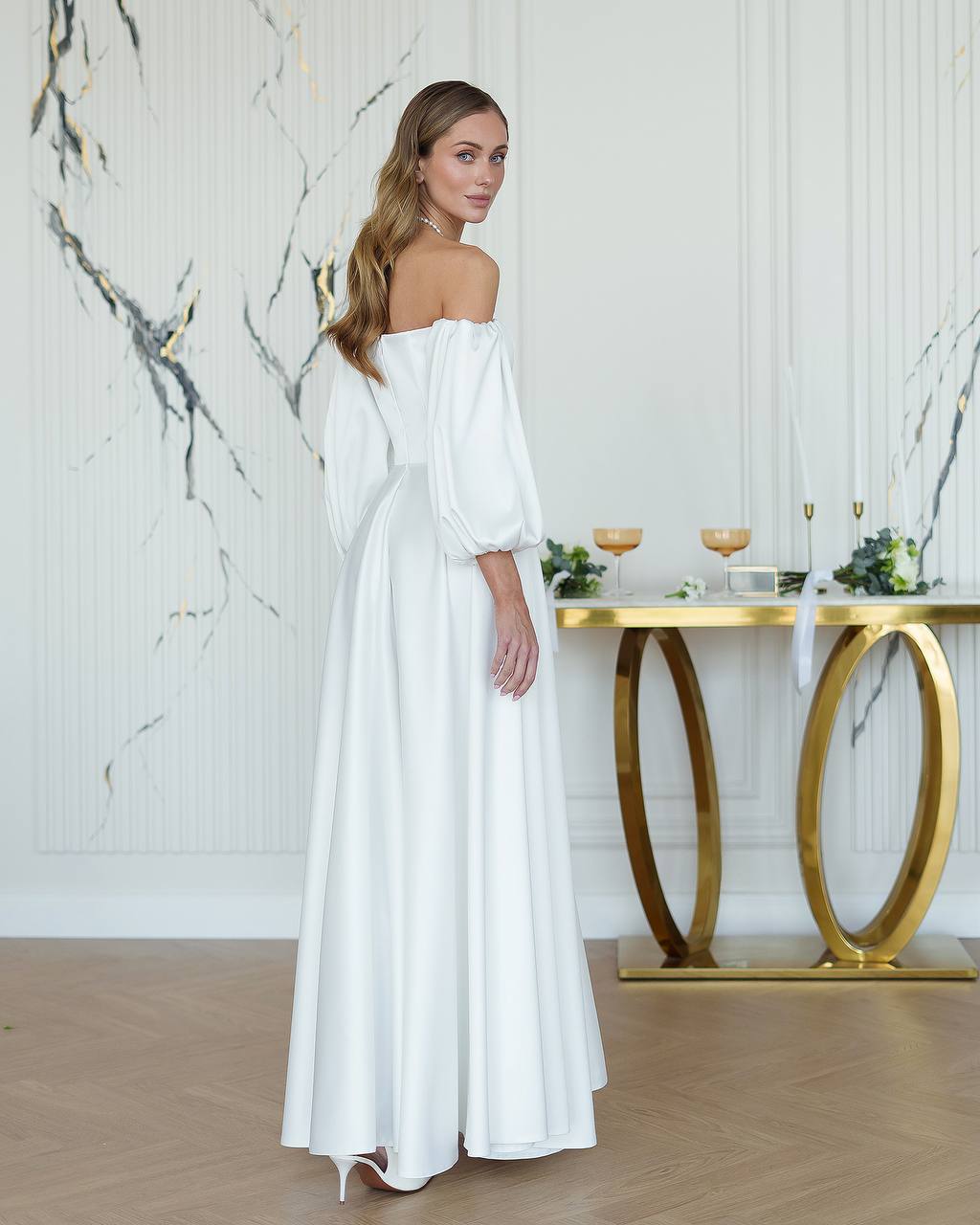 Woman in a white off-shoulder dress standing in a room with marble walls and a gold table.