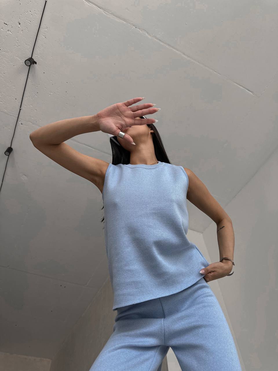 Person wearing a light blue sleeveless top and pants against a plain wall.
