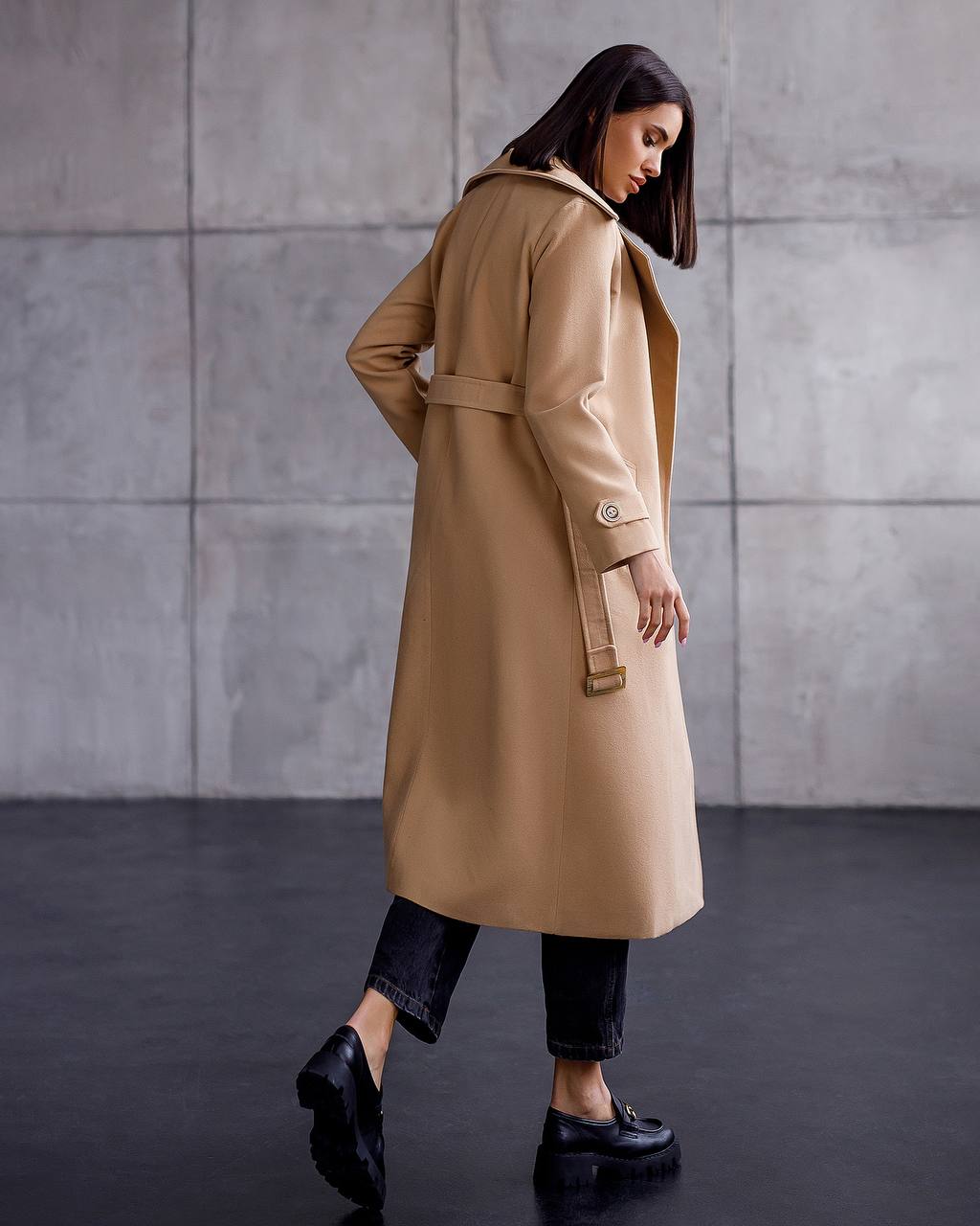 Woman wearing a beige trench coat against a gray wall.