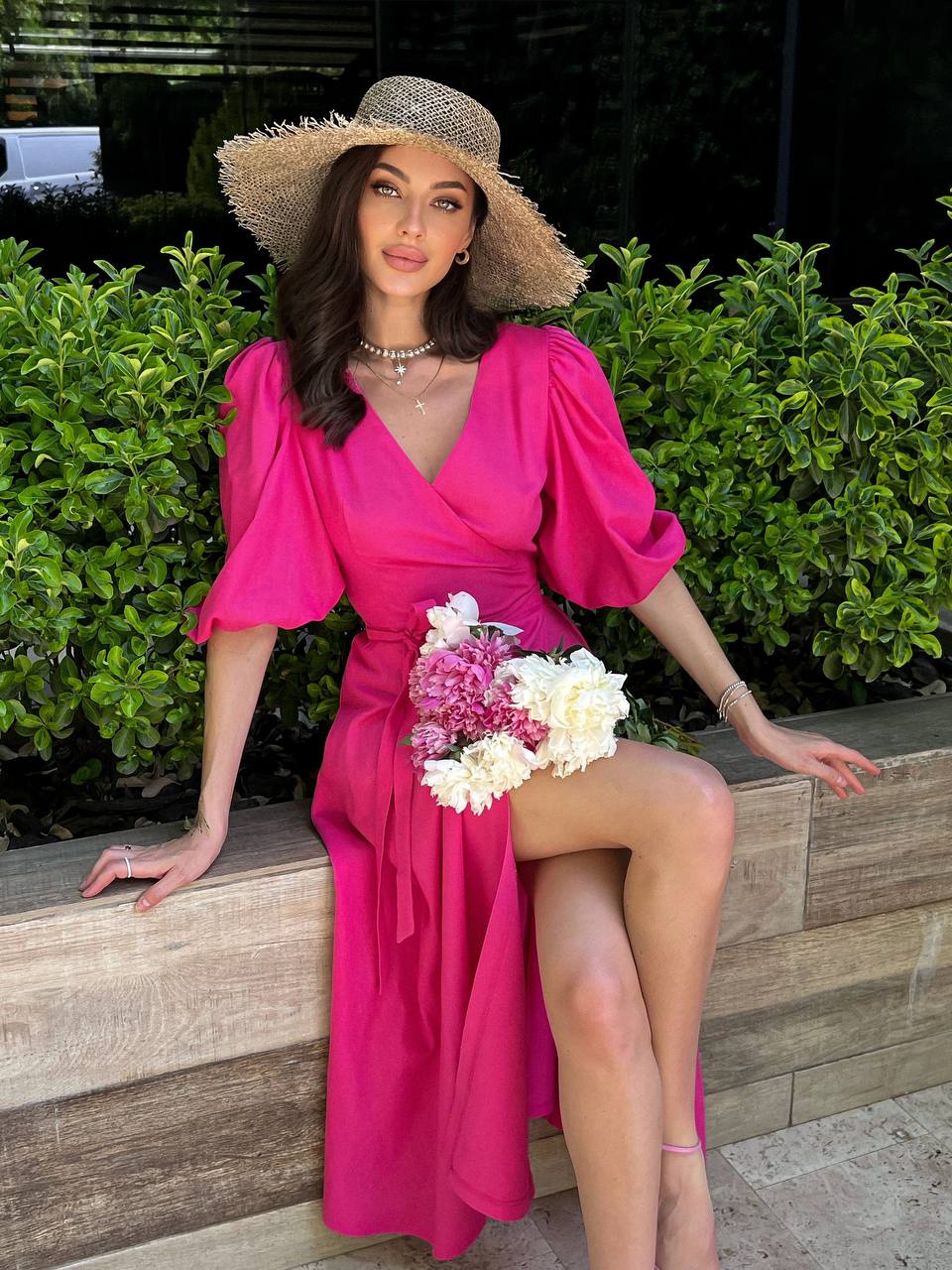 Woman in a bright pink dress and sun hat sitting on a wooden bench with flowers.